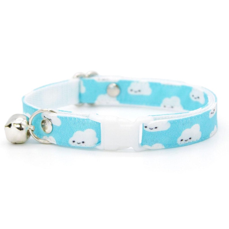 May include: A light blue cat collar with a white cloud pattern and a silver bell. The collar has a white plastic buckle and a metal loop for attaching a leash. The clouds have smiling faces.