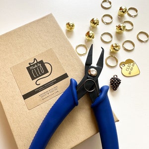 Peut inclure: Une paire de coupe-fils à manches bleues avec une boîte en carton marron en arrière-plan. La boîte porte une étiquette indiquant "Made by Cleo" et "Collars + Accessories for Cats + Little Dogs". Il y a des clochettes dorées, des anneaux de saut et une médaille de chien en forme de cœur dorée avec le texte "Cleo 987-654-3210" sur une surface blanche.