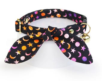 May include: Black cat collar with a bow tie featuring a celestial pattern of stars, moons, and dots in orange, pink, and purple. The collar has a gold-colored buckle and a small gold bell.