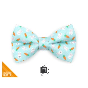 May include: A light blue bow tie with a pattern of white bunnies and orange carrots. The bow tie is made for cats or small dogs. The text "Made by Cleo" is on a white tag.