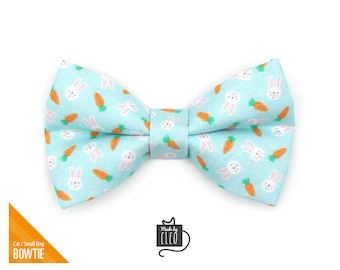 May include: A light blue bow tie with a pattern of white bunnies and orange carrots. The bow tie is made for cats or small dogs. The text "Made by Cleo" is on a white tag.