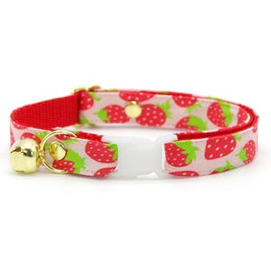 May include: A cat collar with a red strap and a white plastic buckle. The collar features a pattern of red strawberries with green leaves on a light pink background. A gold bell hangs from the collar.