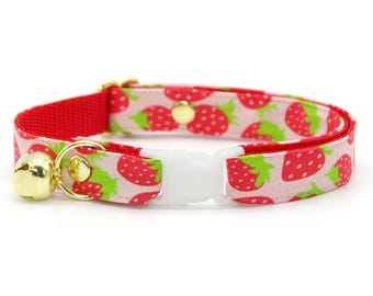 Op de afbeelding: Een kattenhalsband met een rode band en een witte plastic gesp. De halsband heeft een patroon van rode aardbeien met groene bladeren op een lichtroze achtergrond. Een gouden bel hangt aan de halsband.