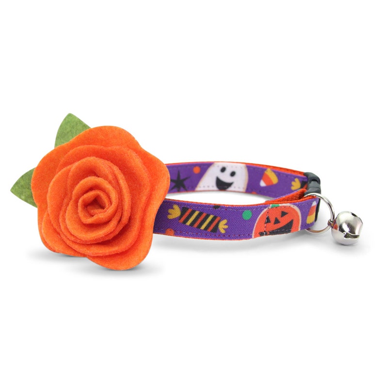 May include: A purple and orange cat collar with a Halloween theme. The collar features a ghost, candy corn, and a jack-o-lantern. A large orange felt flower is attached to the collar.