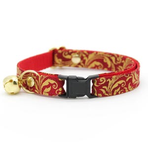 Pode incluir: Coleira vermelha para gatos com sino dourado e fivela de plástico preta. A coleira apresenta um padrão floral dourado e vermelho. O sino é preso com um anel de metal dourado.