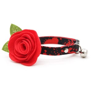 May include: A pet collar featuring a black fabric strap with a red paint splatter pattern. Attached to the collar is a large, layered red felt rose with two green felt leaves. The collar includes a silver metal bell and a black plastic buckle.
