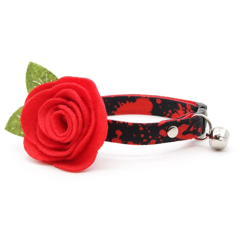 May include: A pet collar featuring a black fabric strap with a red paint splatter pattern. Attached to the collar is a large, layered red felt rose with two green felt leaves. The collar includes a silver metal bell and a black plastic buckle.