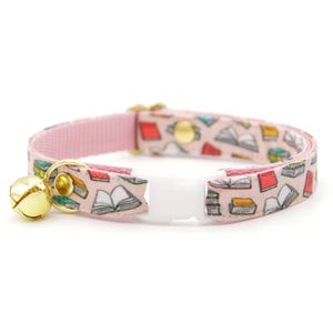 May include: A pink cat collar with a bell and a white plastic buckle. The collar is decorated with a pattern of colorful books.