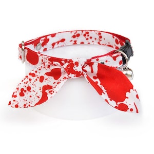 May include: A white cat collar with a red and white bow tie featuring a blood splatter pattern. The collar has a bell and a safety buckle.