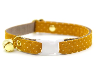 Könnte beinhalten: Senfgelbes Katzenhalsband mit weißen Punkten. Das Halsband hat eine weiße Kunststoffschnalle, eine goldfarbene Glocke und eine goldfarbene Metallschlaufe. Das Halsband ist etwa 1,3 cm breit.