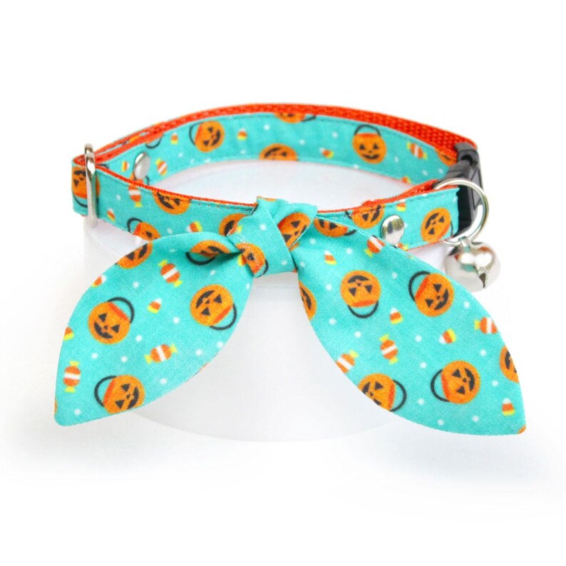 May include: A turquoise blue cat collar with a pumpkin and candy corn pattern. The collar has a bell and a bow tie.