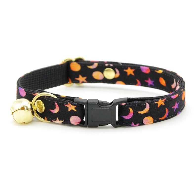 Pu&ograve; includere: Un collare per gatto nero con fibbia di sicurezza e un campanello. Il collare presenta un motivo ripetitivo di lune e stelle rosa, arancioni e gialle su sfondo nero.
