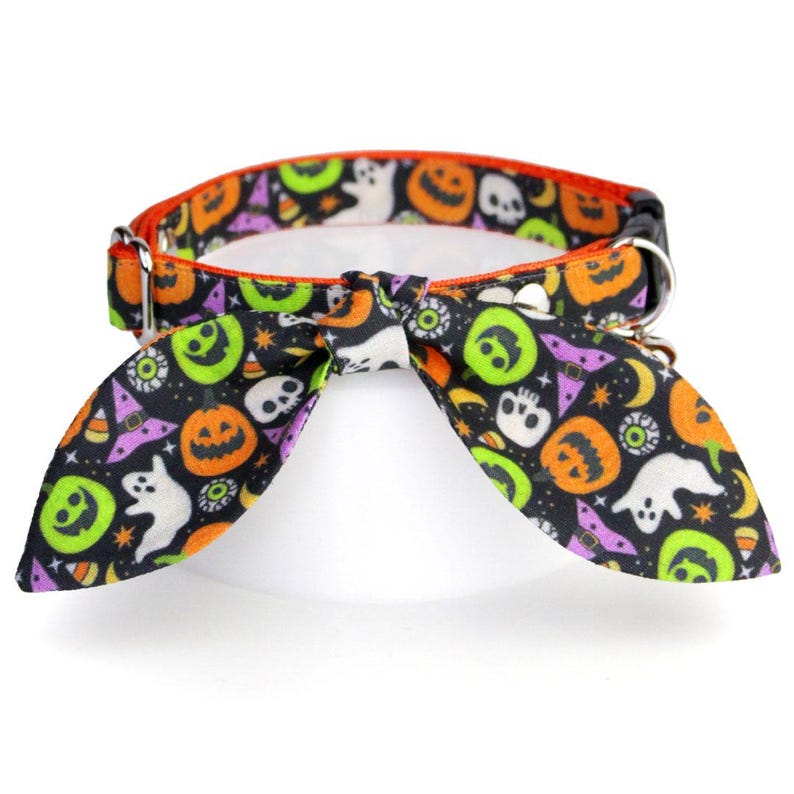 Puede incluir: Un collar para mascotas con tem&aacute;tica de Halloween con un fondo negro y un patr&oacute;n repetido de calabazas, fantasmas y sombreros de bruja. El collar tiene una pajarita y un ribete naranja. Los herrajes met&aacute;licos son plateados.