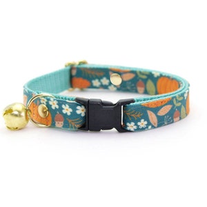 Puede incluir: Un collar para gato azul turquesa con una hebilla de seguridad y una campana. El collar presenta un patrón de calabazas naranjas, bellotas, hojas y flores blancas sobre un fondo verde oscuro.