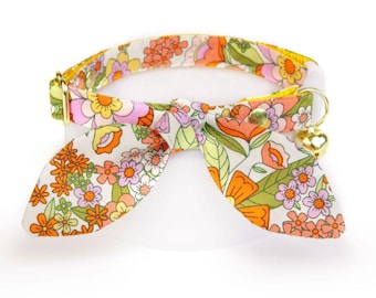 Puede incluir: Un collar de gato blanco con un patrón floral en tonos naranja, rosa y verde. El collar tiene una pajarita y una pequeña campana dorada.