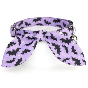 May include: A lavender pet collar with a black bat pattern and a matching bow tie. The collar has a metal buckle and a D-ring for attaching a leash. A small bell hangs from the D-ring. The bow tie is in the shape of two bat wings.