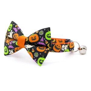 Puede incluir: Un conjunto de collar y pajarita para gatos con tem&aacute;tica de Halloween. La tela negra est&aacute; estampada con calabazas naranjas, fantasmas verdes, sombreros de bruja morados y caramelos blancos. La pajarita tiene un centro naranja y el collar una campana plateada.