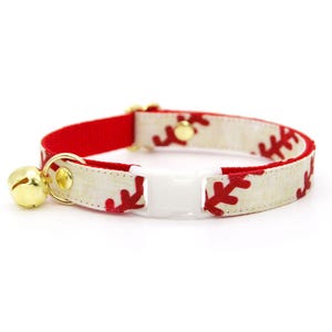 Op de afbeelding: Een kattenhalsband met een honkbalontwerp. De halsband heeft een crèmekleurige stof met rode honkbalstiksels, een rode band en een witte plastic gesp. Een goudkleurige bel hangt aan de halsband.
