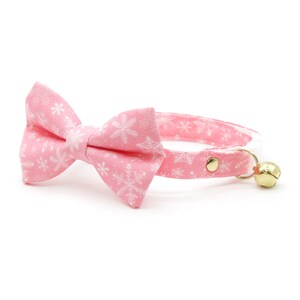 May include: Pink cat collar with a white snowflake pattern and a matching bow tie. The collar has a gold bell.