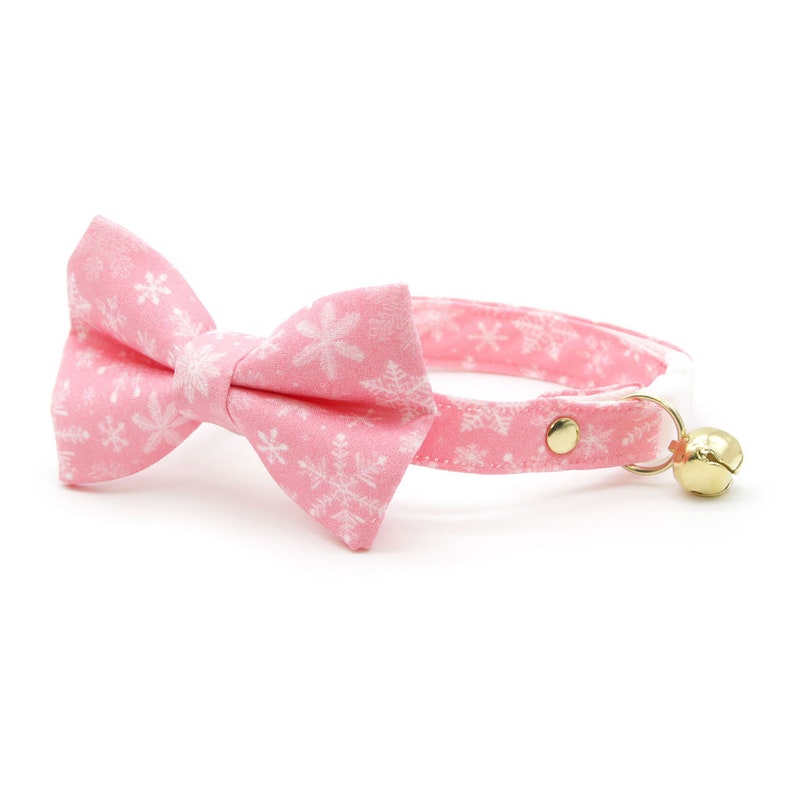 May include: Pink cat collar with a white snowflake pattern and a matching bow tie. The collar has a gold bell.