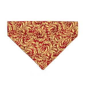 May include: A red and gold patterned dog bandana. The bandana has a repeating gold floral design on a red background. The fabric is folded into a triangle shape, ready to be tied around a dog's neck.