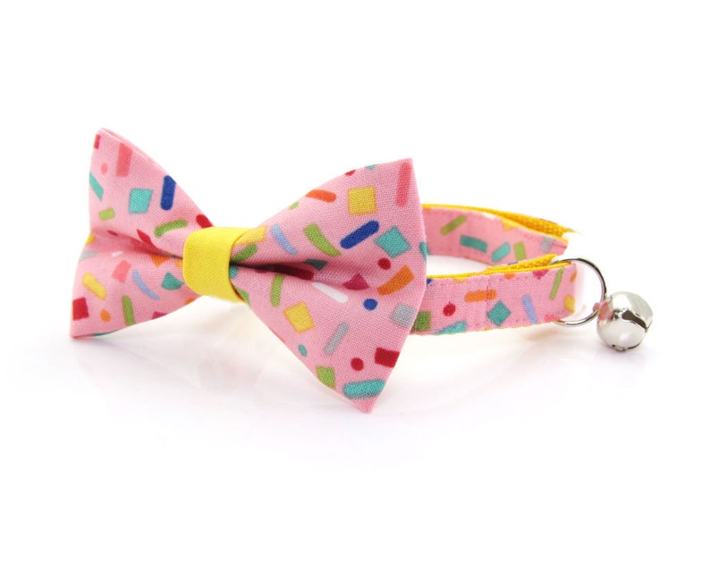 May include: Pink cat collar with a colorful confetti pattern bow tie and a silver bell.