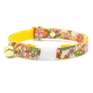 May include: A yellow breakaway cat collar with a floral pattern and a gold bell. The collar has a white plastic buckle.