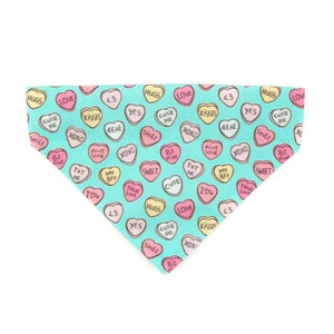 May include: A turquoise dog bandana with a pattern of heart-shaped candies. Each candy has a different message, such as "Hugs," "Love," and "Kisses," in pink, yellow, and white. The bandana is triangular.
