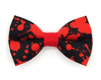 May include: A bow tie featuring a black fabric base with a vibrant red paint splatter pattern. The center of the bow tie is cinched with a solid red fabric band, creating a distinct contrast against the dark background of the accessory.