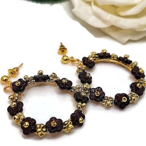 May include: Gold-coloured hoop earrings decorated with dark burgundy flower-shaped beads and smaller gold beads. The earrings have a delicate, handcrafted appearance, with a floral design. The earrings are set against a soft, blurred background.