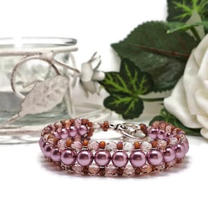May include: A handcrafted bracelet with rows of pink pearls and small brown beads. The bracelet is detailed with clear, faceted beads and a silver clasp. The jewellery is displayed on a white surface with floral accents.