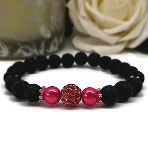 Peut inclure: Un bracelet en pierre de lave noire avec des détails roses. Le bracelet présente un groupe central de petites perles roses scintillantes, flanqué de deux grosses perles roses rondes. Le bracelet est composé de pierres de lave noires.