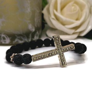 May include: A black lava bead bracelet with a silver-coloured cross pendant adorned with small, clear crystals. The cross is the central element, set against the dark beads. A white rose and a candle are in the background.