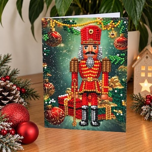 May include: A diamond painting greeting card featuring a central nutcracker figure in a red and gold uniform, standing before wrapped gifts. The background displays a festive green scene with hanging ornaments, pine branches, and golden accents.