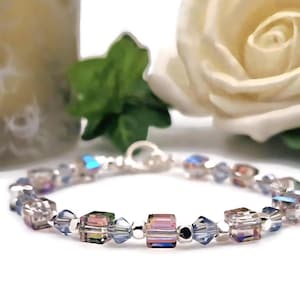 May include: An elegant bracelet with iridescent crystal beads in blue, pink, and clear. The bracelet has a silver clasp. It is displayed on a white surface with a blurred background of a candle and a white rose.