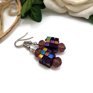 May include: Handmade earrings with a cube design, featuring iridescent, purple, and brown beads. The earrings have silver-coloured hooks. The earrings are displayed on a white surface.