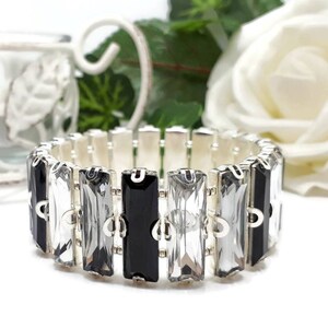 May include: A silver bracelet featuring rectangular black, clear, and grey crystal-like stones. The stones are linked by silver metal, forming a flexible, stretchable design. The bracelet is presented on a white surface.