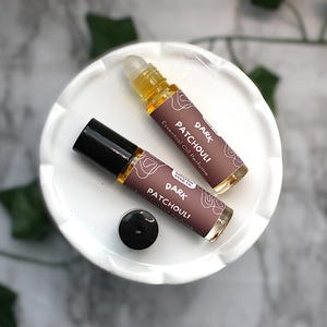 May include: Two glass rollerball perfume bottles containing amber-colored liquid. Each bottle features a brown label with white text that reads DARK PATCHOULI Essential Oil Perfume and a small black cap, with one cap removed and placed nearby.