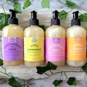 May include: Four clear plastic bottles of moisturizing hand soap with black pump dispensers. Each 8 fl oz bottle features a colorful label with the text Olive Blossom Soap Co. and scents including French Lavender & Honey, Citrus Lemongrass, Black Raspberry Vanilla, and Orange Blossom.