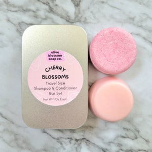 May include: A travel-size shampoo and conditioner bar set in a silver tin. The set includes two pink bars and a tin with a pink label that reads "Cherry Blossoms" and "Travel Size Shampoo & Conditioner Bar Set". Each bar weighs 1 oz.