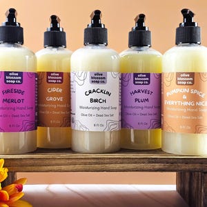 May include: Five clear plastic bottles of moisturizing hand soap with black pump dispensers. Each bottle features a label with the Olive Blossom Soap Co. logo, scent name, and 8 fl oz volume. Scents include Fireside Merlot, Cider Grove, Cracklin Birch, Harvest Plum, and Pumpkin Spice & Everything Nice.