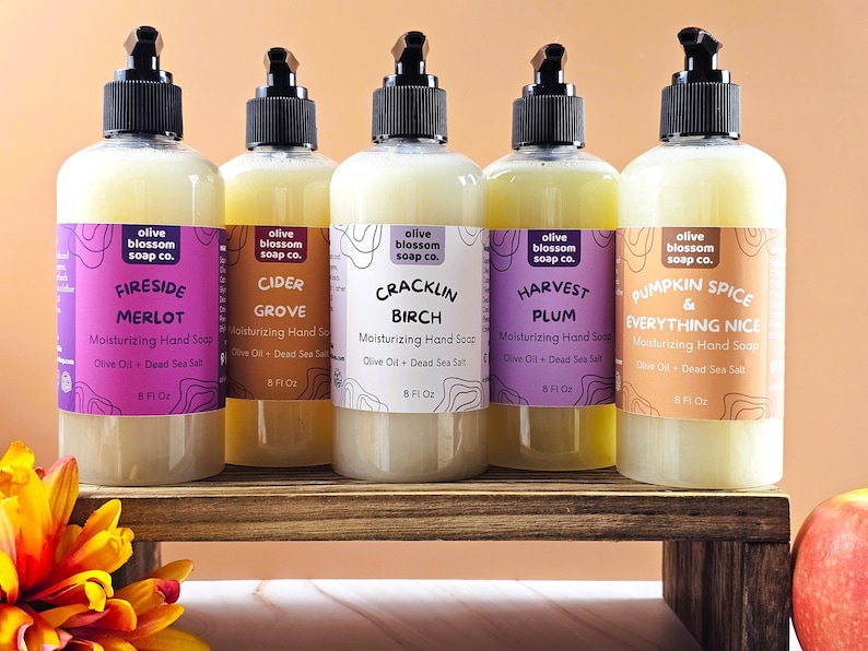 May include: Five clear plastic bottles of moisturizing hand soap with black pump dispensers. Each bottle features a label with the Olive Blossom Soap Co. logo, scent name, and 8 fl oz volume. Scents include Fireside Merlot, Cider Grove, Cracklin Birch, Harvest Plum, and Pumpkin Spice & Everything Nice.