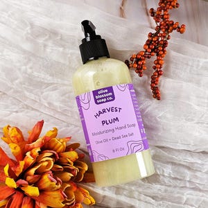 May include: A clear plastic bottle of Harvest Plum moisturizing hand soap with a black pump dispenser. The purple label features the text olive blossom soap co., Harvest Plum, Moisturizing Hand Soap, Olive Oil + Dead Sea Salt, and 8 Fl Oz.