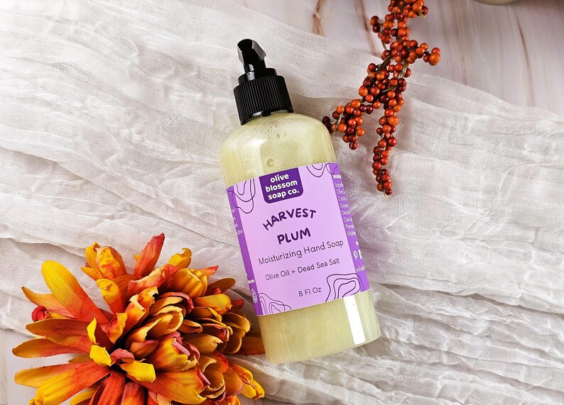 May include: A clear plastic bottle of Harvest Plum moisturizing hand soap with a black pump dispenser. The purple label features the text olive blossom soap co., Harvest Plum, Moisturizing Hand Soap, Olive Oil + Dead Sea Salt, and 8 Fl Oz.