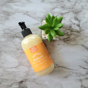 May include: A clear plastic bottle of Orange Blossom moisturizing hand soap with a black pump dispenser. The orange label features the text olive blossom soap co., ORANGE BLOSSOM, Moisturizing Hand Soap, Olive Oil + Dead Sea Salt, 8 Fl Oz.
