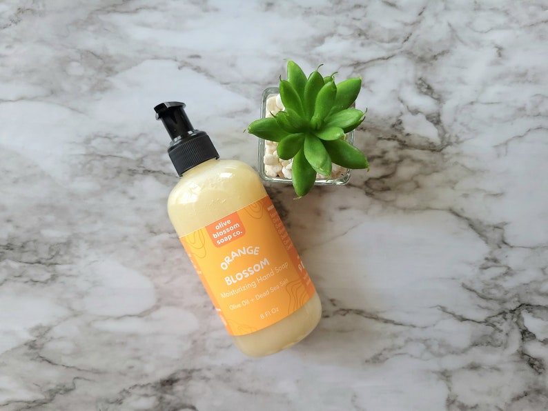 May include: A clear plastic bottle of Orange Blossom moisturizing hand soap with a black pump dispenser. The orange label features the text olive blossom soap co., ORANGE BLOSSOM, Moisturizing Hand Soap, Olive Oil + Dead Sea Salt, 8 Fl Oz.