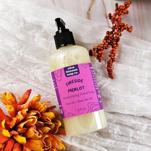 May include: A clear plastic bottle of moisturizing hand soap with a black pump dispenser. The label is bright purple with white text reading Olive Blossom Soap Co., Fireside Merlot, Moisturizing Hand Soap, Olive Oil + Dead Sea Salt, 8 Fl Oz.