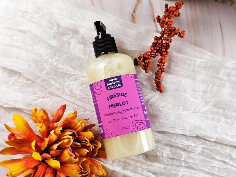 May include: A clear plastic bottle of moisturizing hand soap with a black pump dispenser. The label is bright purple with white text reading Olive Blossom Soap Co., Fireside Merlot, Moisturizing Hand Soap, Olive Oil + Dead Sea Salt, 8 Fl Oz.