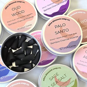May include: A collection of round metal tins containing bamboo charcoal incense cones. Each lid features the Olive Blossom Soap Co. brand name, scent names like Oud Wood and Palo Santo, and the text 15 1-inch cones.