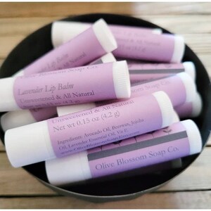 May include: A selection of lavender lip balm tubes in a black bowl. Each tube features a white base and a light purple label with the text "Lavender Lip Balm, Unsweetened & All Natural". The net weight is 4.2 grams.